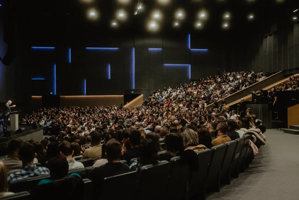 A packed modern auditorium with a speaker on stage addressing the audience.