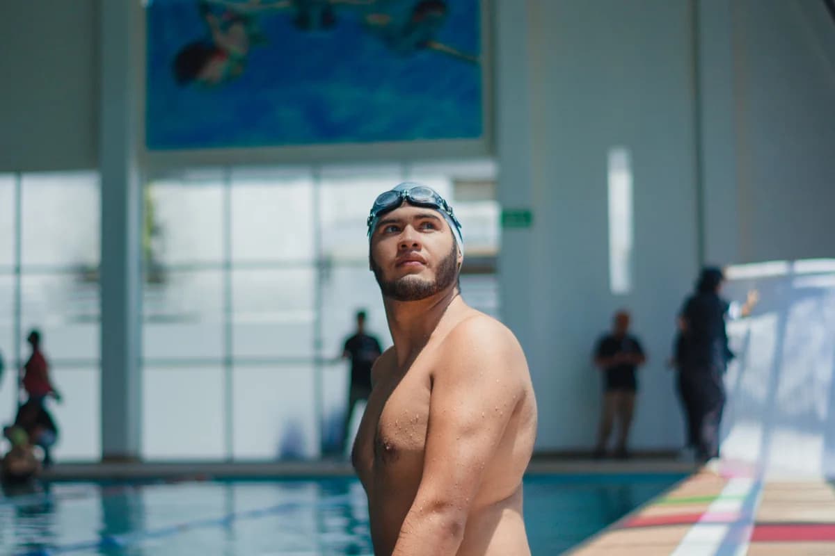 A swimmer in goggles stands by an indoor pool, looking up in concentration.