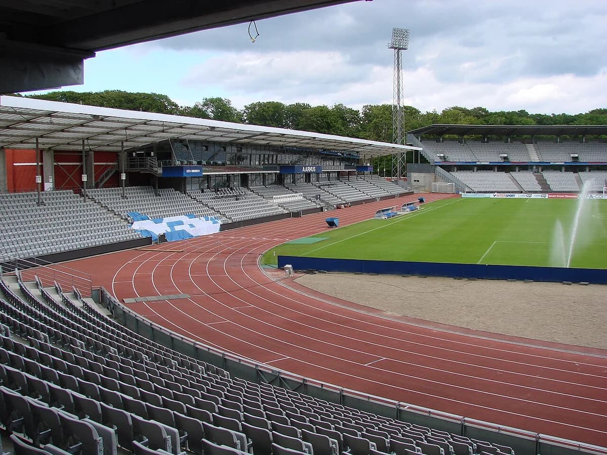 Empty Aarhus Stadium with gray seats, a green pitch, and a red running track.