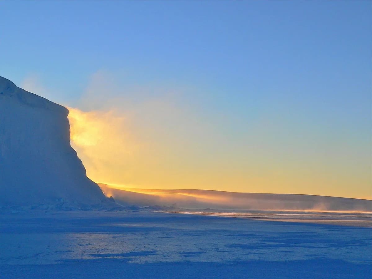 Sunrise over Antarctic icebergs with a clear blue sky and glowing horizon.
