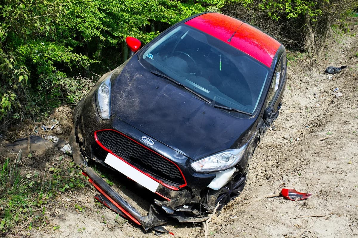 A damaged black and red Ford car sits in a ditch beside a dirt road.