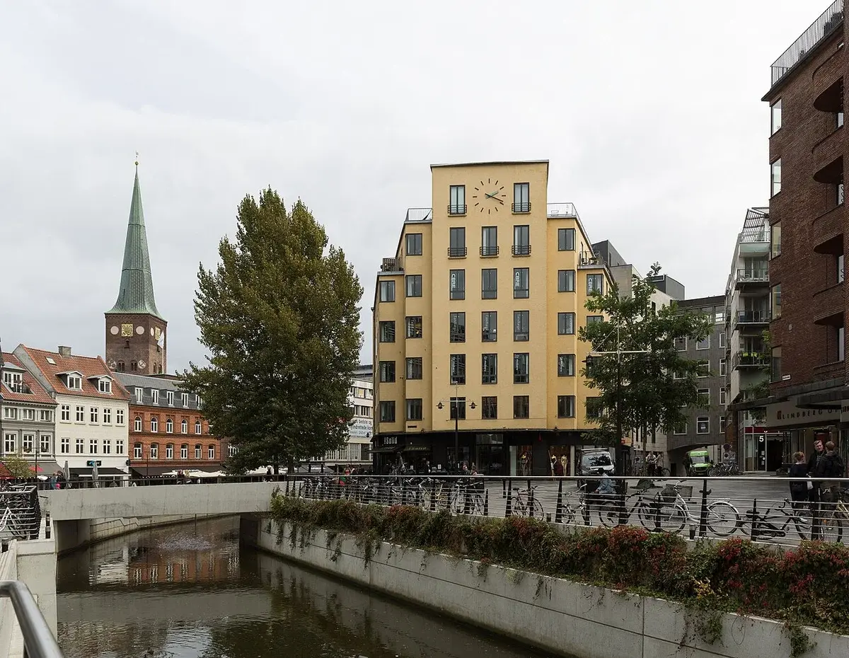 Urban river scene in Aarhus with buildings, bicycles, pedestrians, and a church spire.