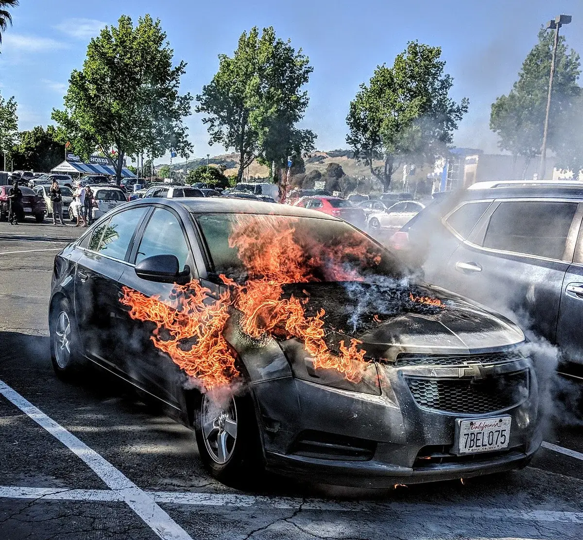 A car on fire in a parking lot with smoke rising around it.