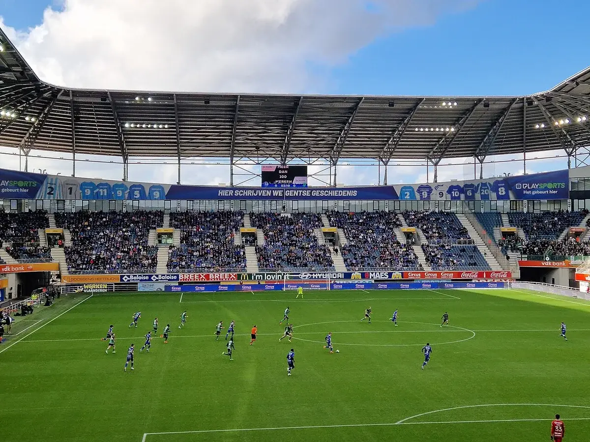A football match in progress at KAA Gent's stadium with a full crowd in the stands.
