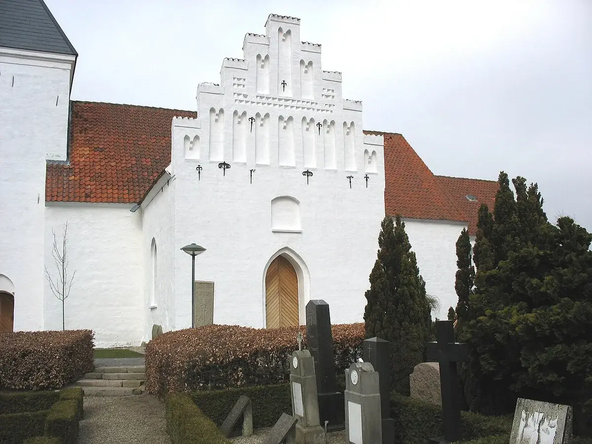 Side view of a white church with a red tile roof beside gravestones and trimmed hedges.