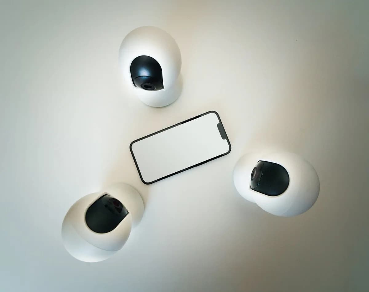 A smartphone surrounded by three security cameras on a plain light background.