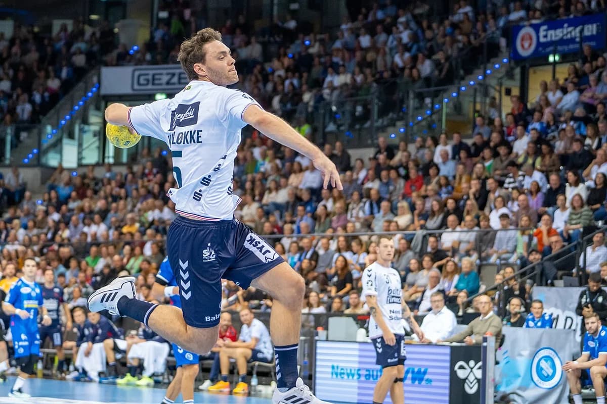 A handball player in a white jersey jumps to throw the ball during a match in a packed arena.